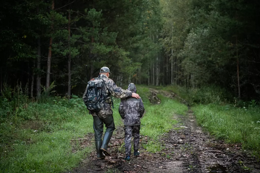 Hunters with hunting equipment going away through rural field towards forest at sunset during hunting season in countryside