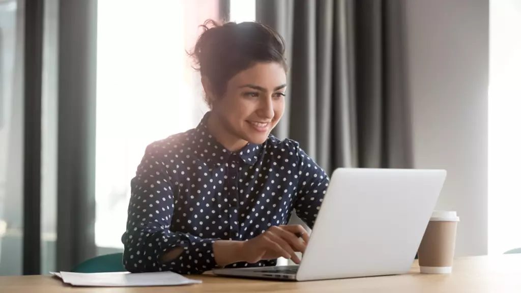 Happy young indian business woman entrepreneur using computer looking at screen working in internet sit at office desk, smiling hindu female professional employee typing email on laptop at workplace